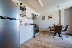A modern kitchen with white cabinets, a stainless steel refrigerator, a microwave, and an oven. There is a dining area with a table and chairs.