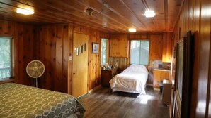 Una habitación tipo cabaña con paredes y pisos de madera, dos camas, un ventilador y una pequeña zona de escritorio.