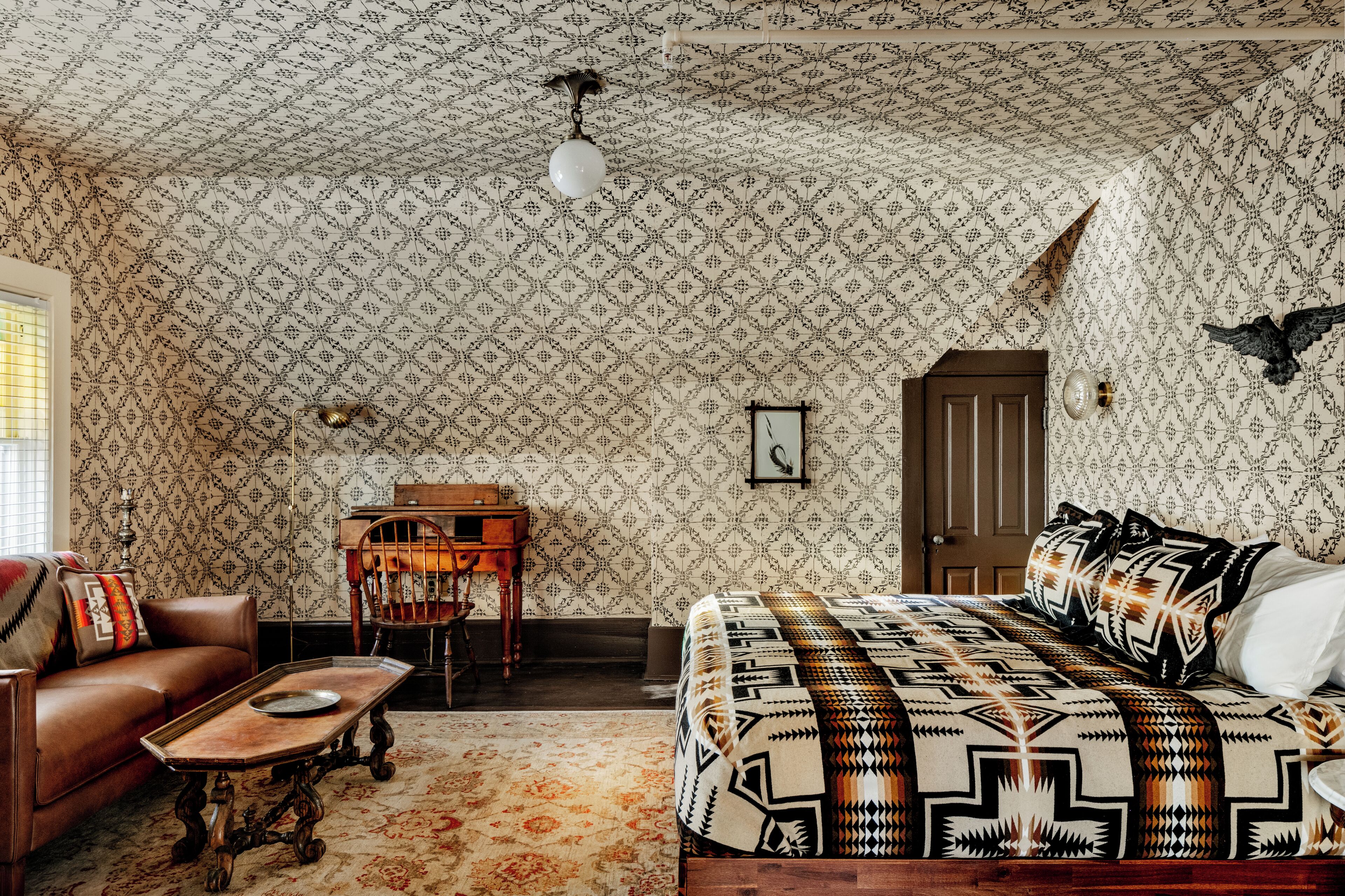 Una habitación con papel tapiz estampado, una cama con colcha geométrica, una silla de madera, una mesita y una lámpara.