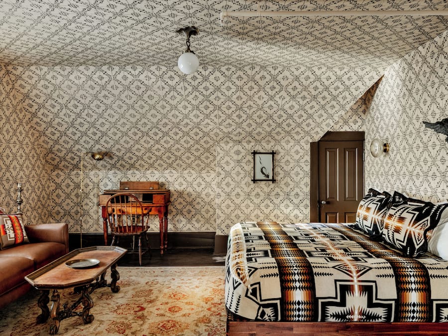 Una habitación con papel tapiz estampado, una cama con colcha geométrica, una silla de madera, una mesita y una lámpara.