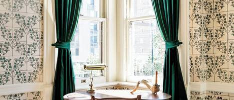 A corner room with patterned wallpaper, a wooden desk with drawers, a chair, a window with green curtains, and a decorative item on the windowsill.