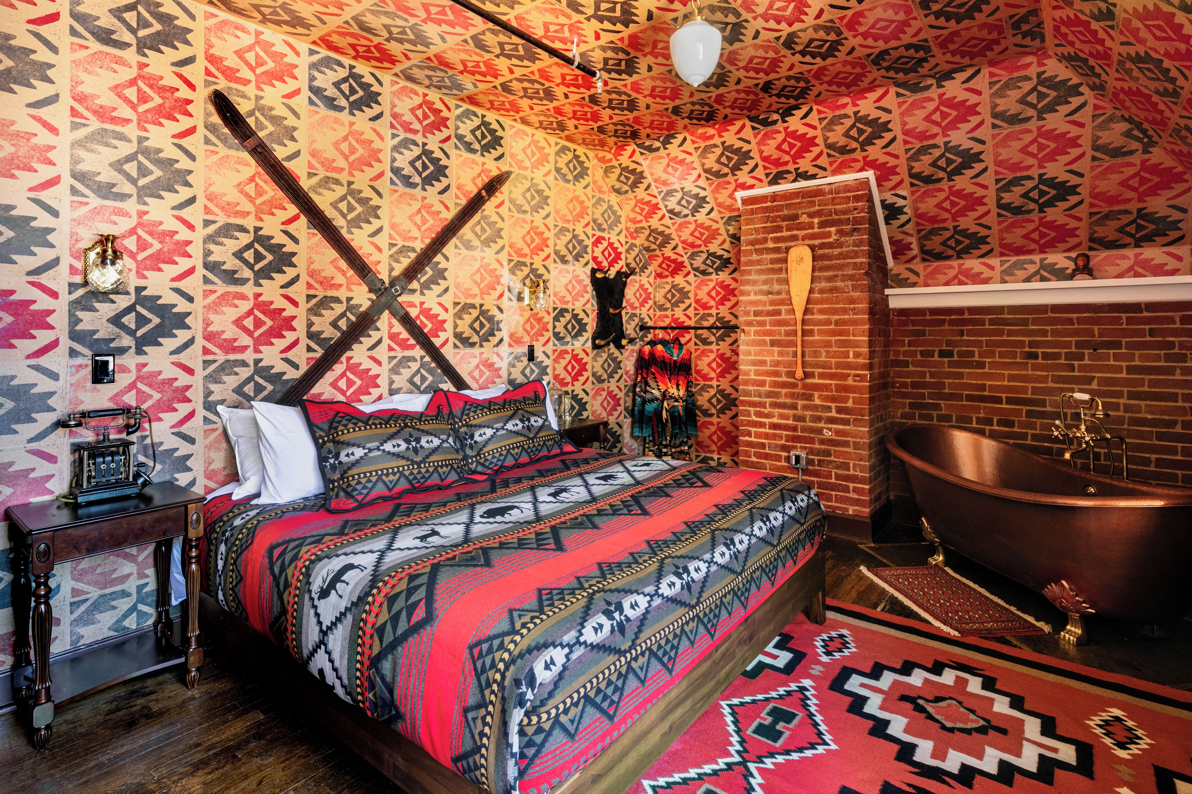 A room with a patterned wall, a bed with a red and black bedspread, a wooden nightstand, a vintage radio, a brick wall, and a freestanding bathtub.