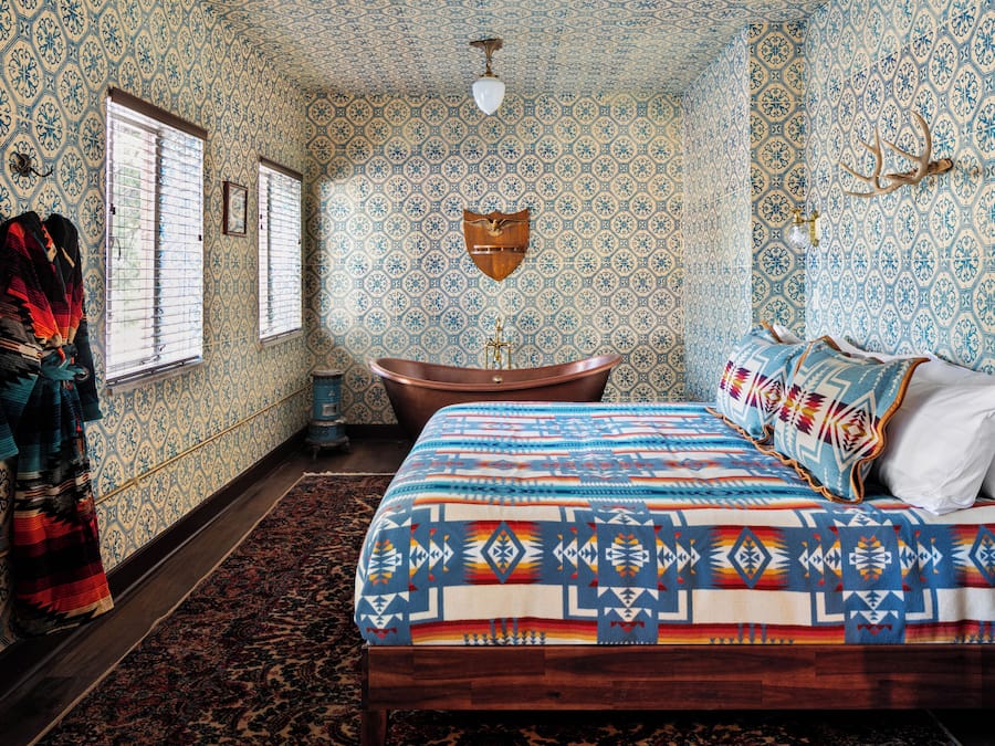Una habitación con papel tapiz estampado, una cama con colcha estampada, un banco de madera y una bañera.
