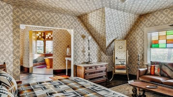 Una habitación con papel tapiz estampado, una cama con colcha de diseño geométrico, un aparador de madera, un espejo y un sofá de cuero.