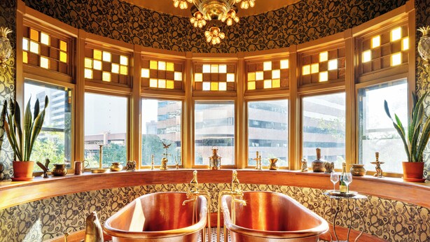 Une salle de bain d’angle avec deux vasques en cuivre, des encadrements de fenêtres décoratifs et un luminaire ornemental au plafond.