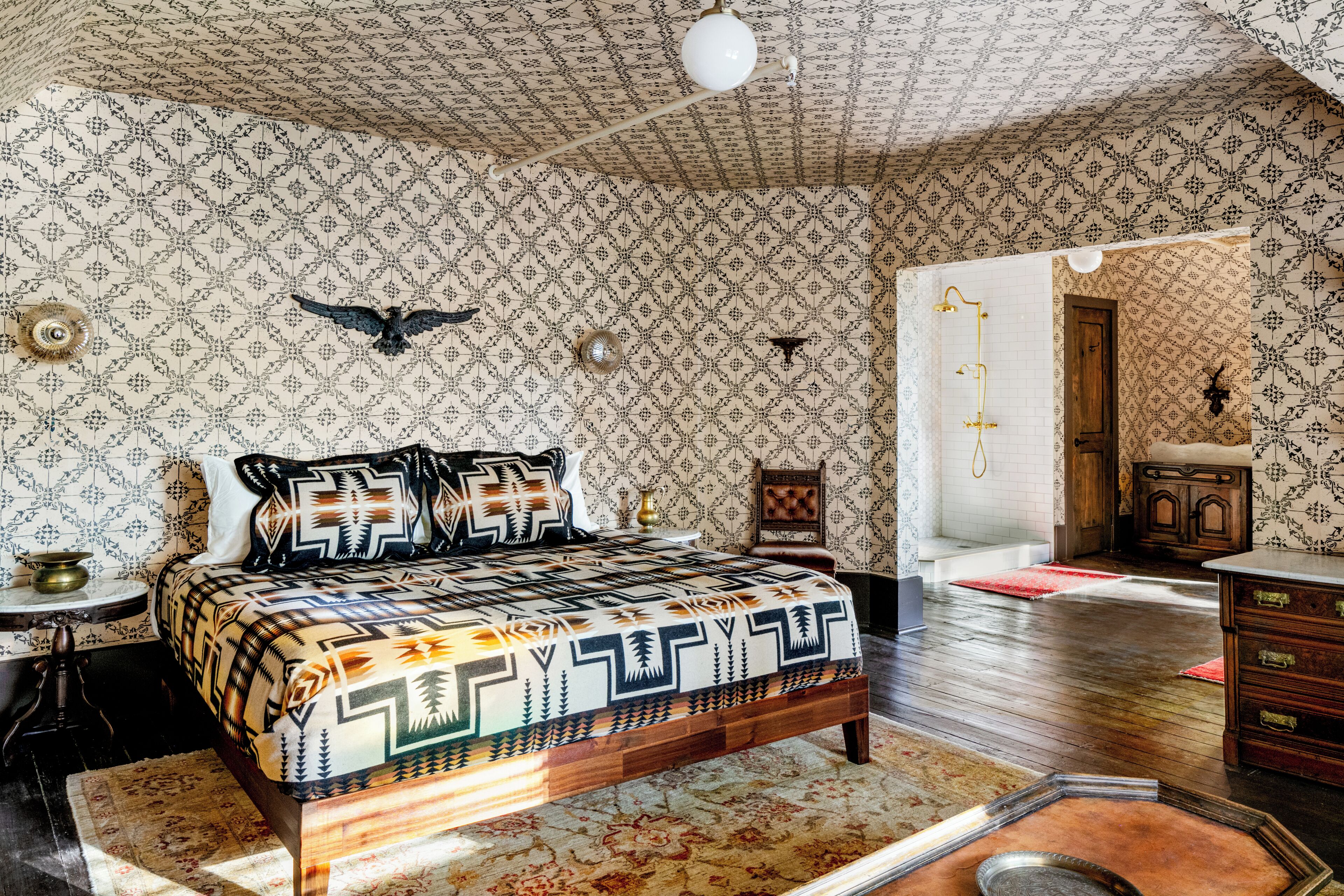 A room with patterned wallpaper, a bed with a geometric bedspread, a wooden dresser, and a red rug.