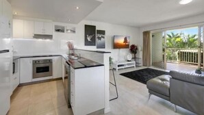 A modern kitchen with white cabinetry, a central island, and a dining area with a grey sofa. - Lennox Beach Resort (Lennox Head)