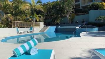 Piscina com espreguiçadeiras e toalha, rodeada por palmeiras e um edifício ao fundo.
