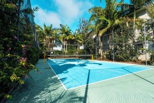 Una cancha de tenis al aire libre rodeada de densa vegetación tropical y un edificio al fondo.