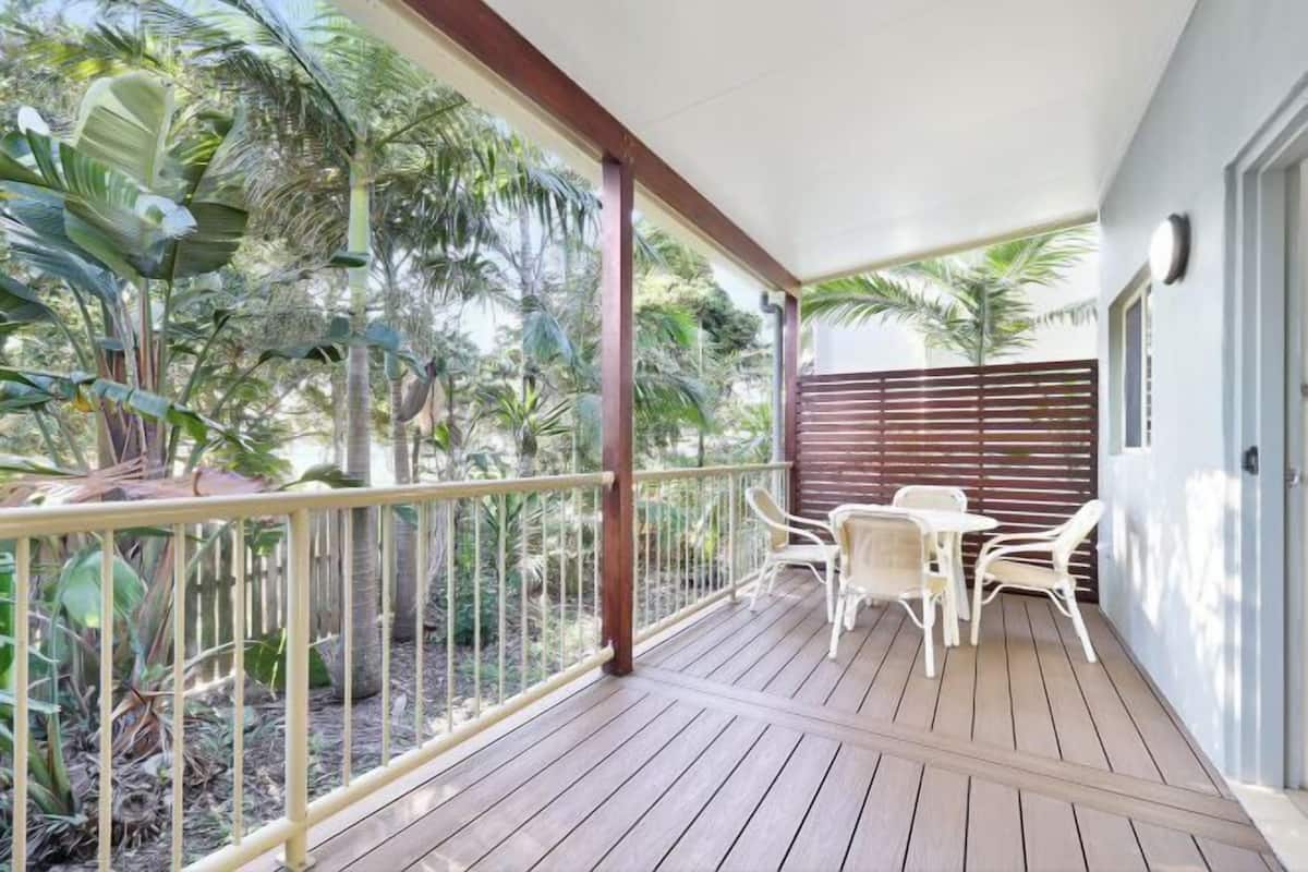 A covered outdoor area with a wooden deck, white chairs, and a view of tropical plants.