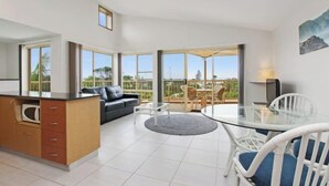 A modern kitchen with a built-in microwave, a dining area with a glass table and white chairs, and a sofa with a view of the outdoors. - Lennox Beach Resort (Lennox Head)
