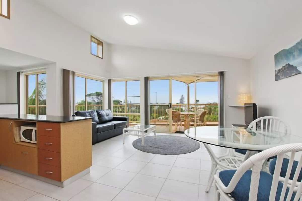 A modern kitchen with a built-in microwave, a dining area with a glass table and white chairs, and a sofa with a view of the outdoors.