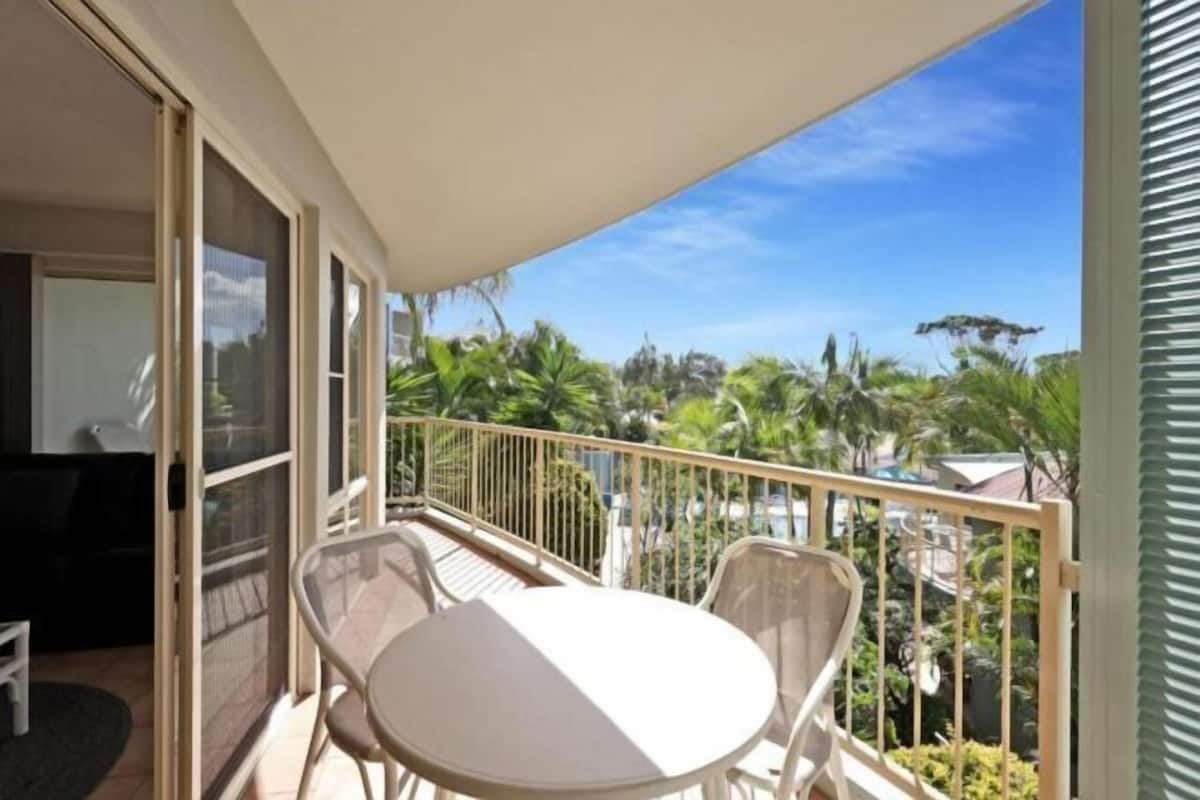A balcony with a table and chairs, a sliding glass door, and a view of palm trees and buildings.