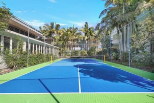 Pista de tenis al aire libre con red, rodeada de vegetación y un edificio.