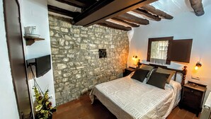 A bedroom with a stone wall, a bed with patterned bedding, a nightstand with a lamp, and a window with a view. - CAN PINÓS DE GUIMERÀ  (Guimerà)