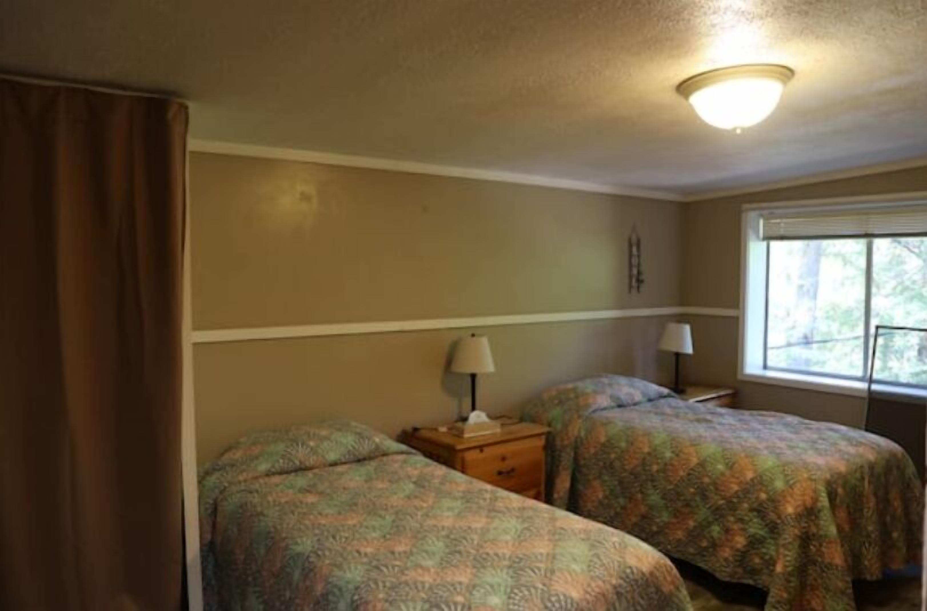 Quarto com duas camas, janela com persianas, luminária de teto e mesa de cabeceira com abajur.