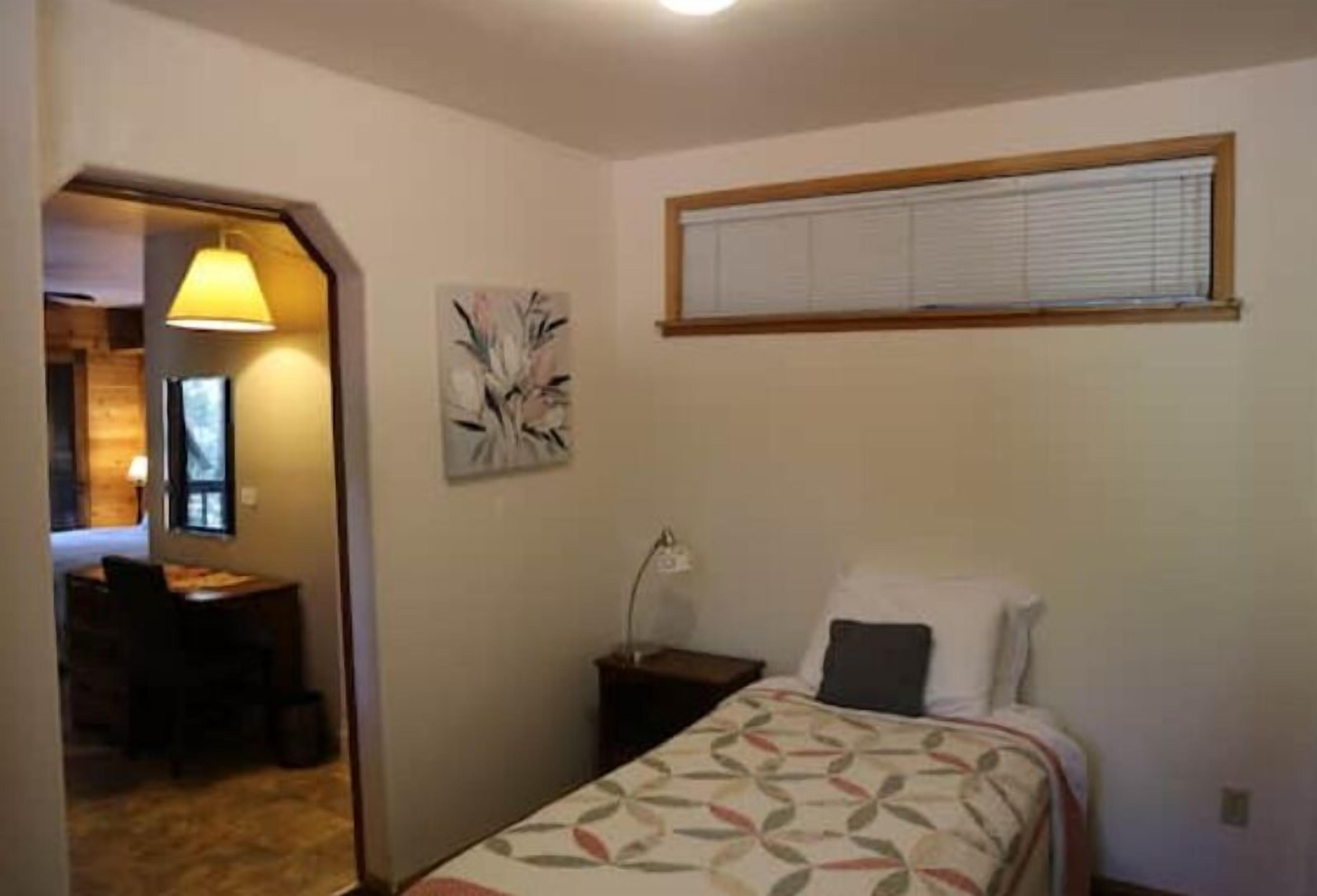 A bedroom with a bed, a desk, and a chair. There is a window with blinds and a painting on the wall.