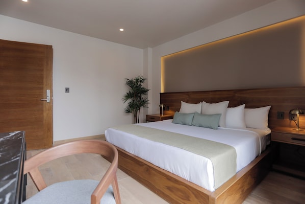 A modern hotel room with a wooden bed frame, white bedding, a bedside table with a lamp, and a potted plant.