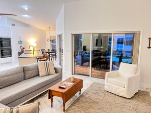 Una sala de estar moderna con un sofá gris, un sillón blanco y una mesa de centro de madera. Unos puertas de cristal dan acceso a un patio con mobiliario exterior.