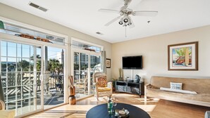 Un vaste salon doté d’une grande porte-fenêtre coulissante donnant sur un balcon, d’un ventilateur de plafond, d’un téléviseur à écran plat et d’un canapé agrémenté d’un coussin sur lequel est inscrit « Folly Beach ».