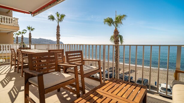 A balcony with wooden furniture, a view of the beach, and palm trees. - Stunning apartment in Almuñecar (Almuñecar)