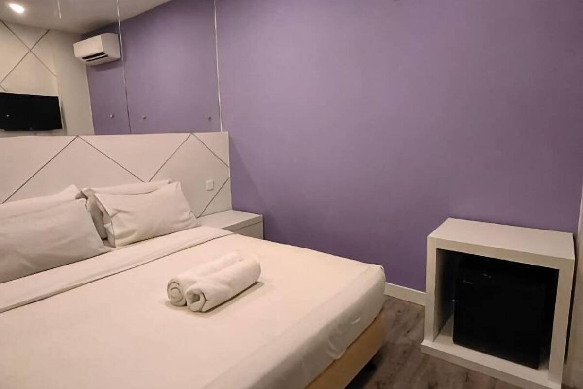 Cama bem arrumada com lençóis brancos e uma toalha enrolada, mesa de cabeceira branca e uma televisão em um suporte.