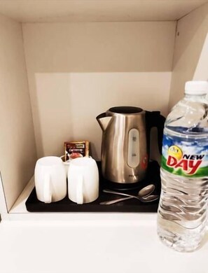 Una kitchenette con un hervidor de acero inoxidable, dos tazas blancas, una botella de agua y una pequeña caja de leche para café.