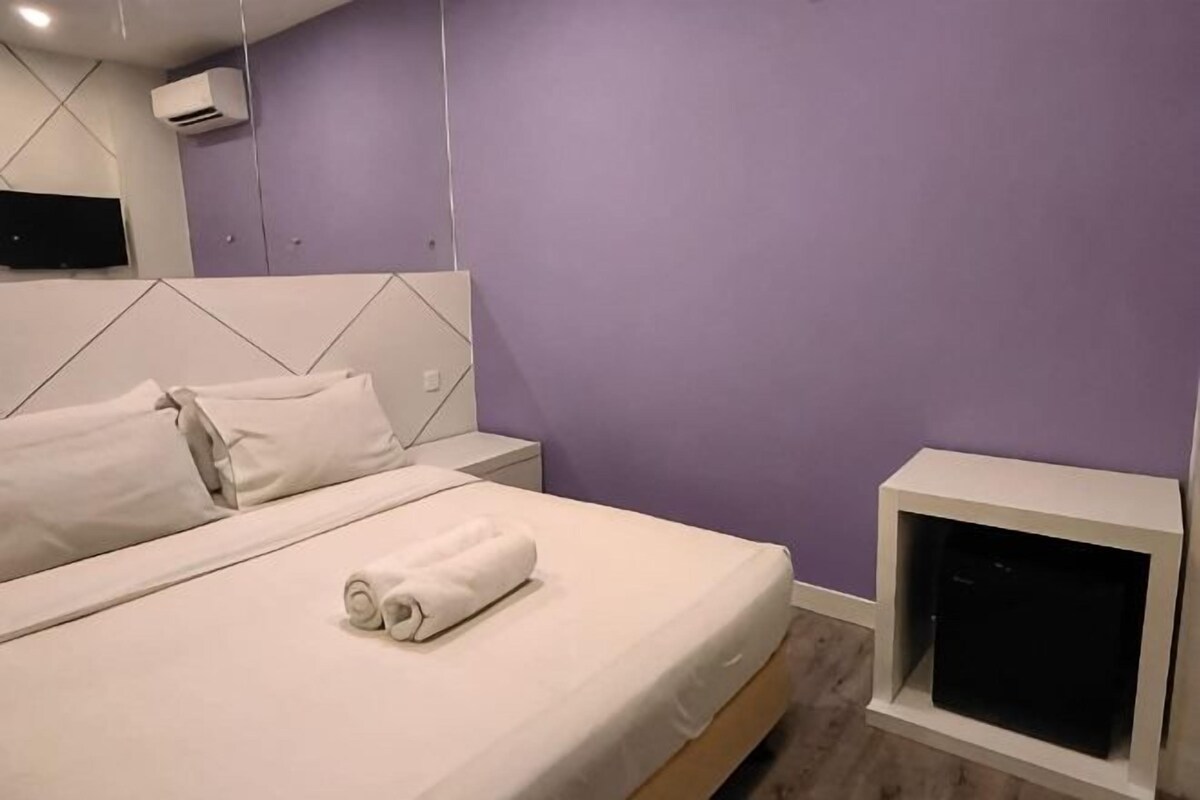 A neatly made bed with white linens and a rolled towel, a white bedside table, and a television on a stand.