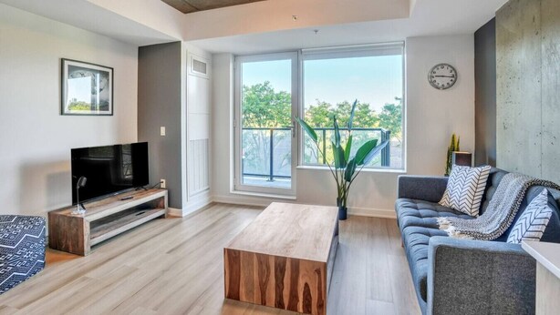 A modern living room with a flat-screen TV, a wooden coffee table, a gray sofa with patterned cushions, and a large window offering a view of trees. - Chic Urban 1BR Apt with Balcony and Skyline Views (Ottawa)