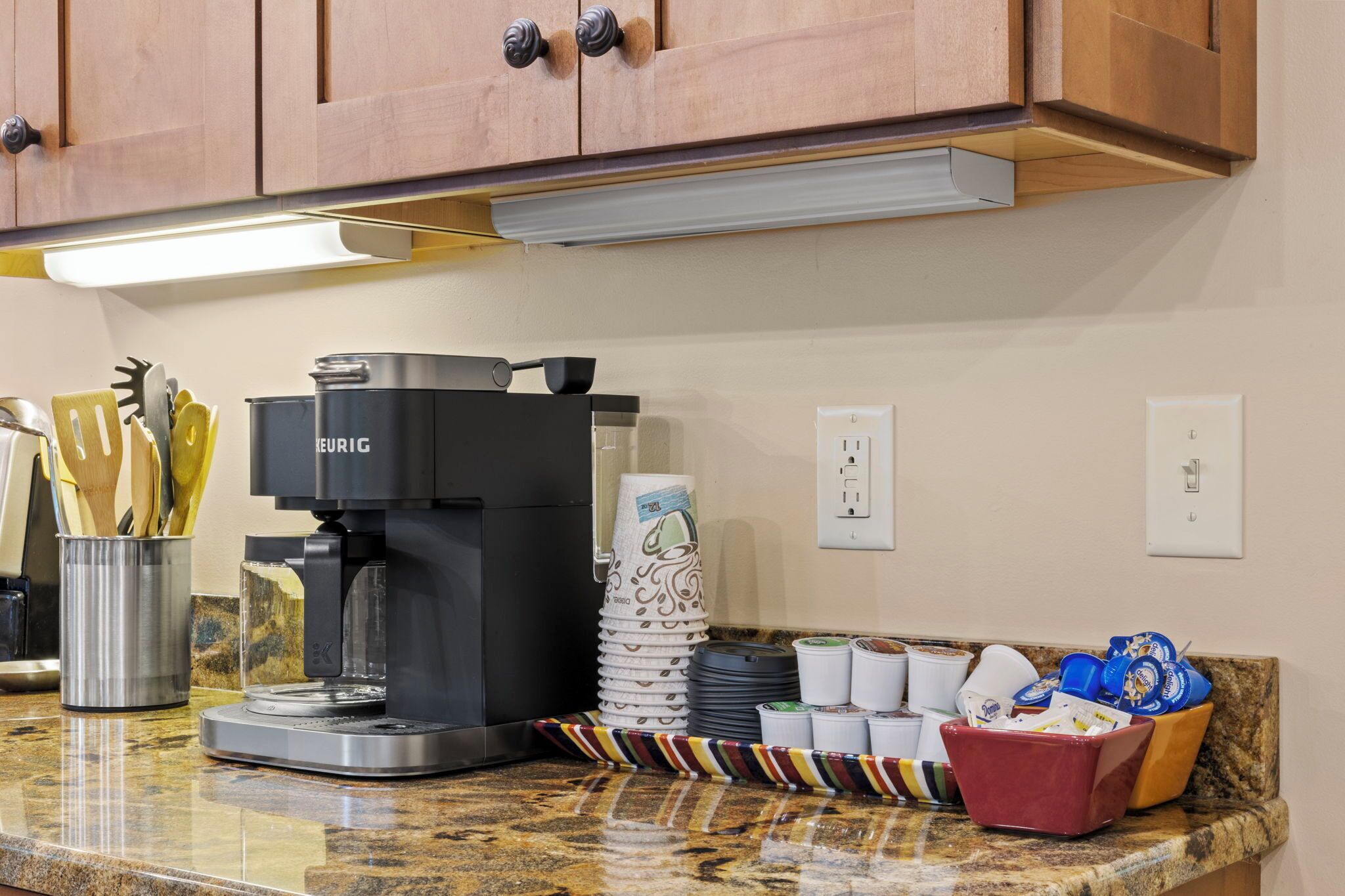 A kitchen with a coffee maker, stacked paper cups, and a utensil holder.