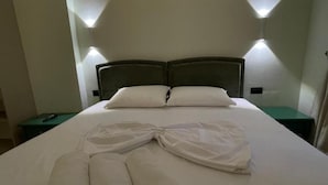 Een net opgemaakt bed met witte lakens en handdoeken, twee nachtkastjes en wandlampen.
