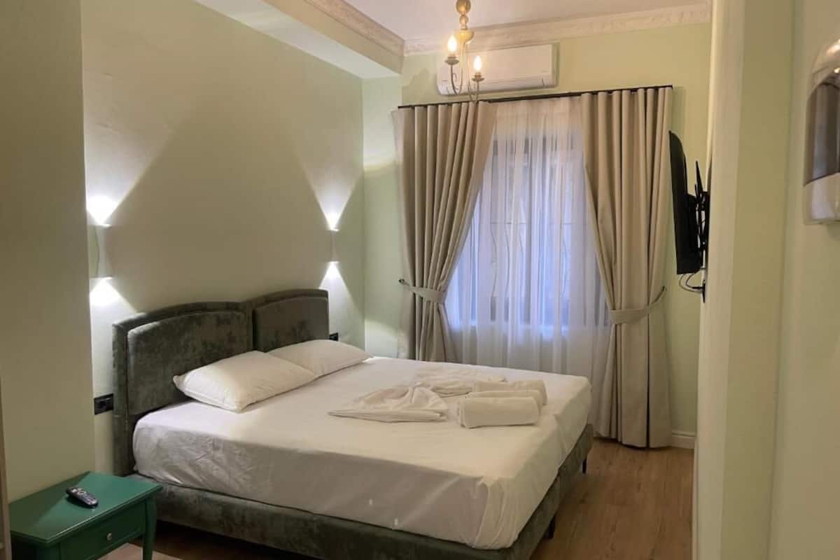 Un lit bien fait, avec du linge de lit blanc et deux serviettes, une table de chevet avec une lampe et un téléviseur fixé au mur.
