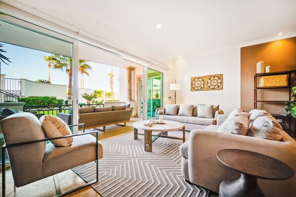 Un salon moderne avec un canapé, des fauteuils et une table basse. Une grande fenêtre offre une vue sur les palmiers et donne accès à un balcon.