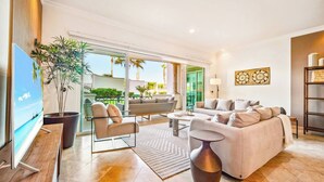 A spacious living room with a large sofa, a coffee table, and a TV. There is a large window with a view of the outdoors.