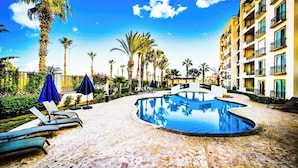 A swimming pool with lounge chairs and umbrellas, surrounded by palm trees and a multi-story building.
