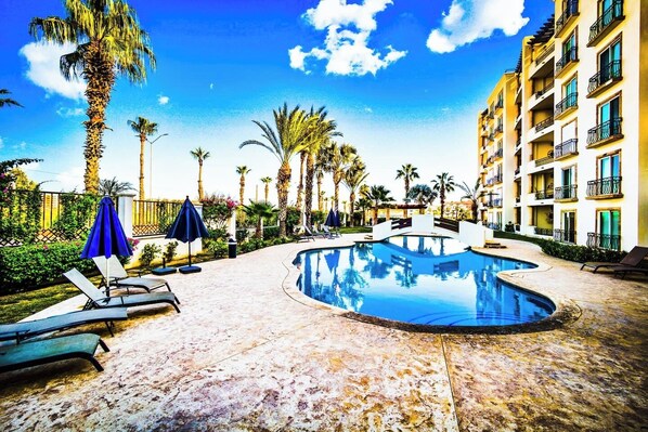 A swimming pool with lounge chairs and umbrellas, surrounded by palm trees and a multi-story building.