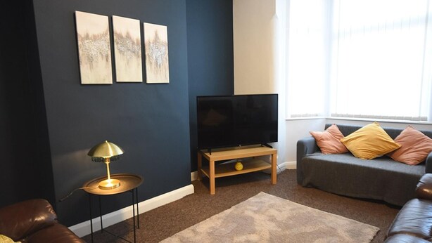 Ein Wohnzimmer mit dunkelblauer Wandgestaltung, einem Flachbildfernseher auf einem Holzständer, einem braunen Ledersofa mit Kissen, einem kleinen runden Tisch mit einer Lampe und einem großen Fenster mit Jalousien.