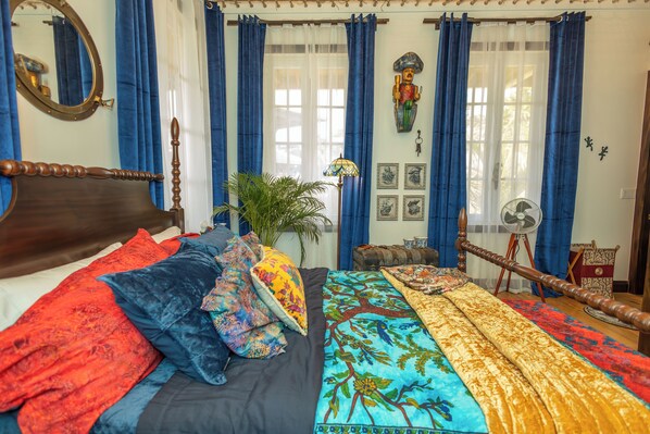Ein Schlafzimmer mit einem hölzernen Bett, blauen Vorhängen, einem Ventilator und einer Wanddekoration.