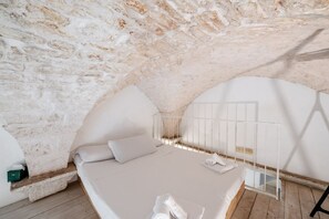 Quarto com teto de pedra, cama com lençóis brancos, uma pequena mesa e janela com grades de metal.