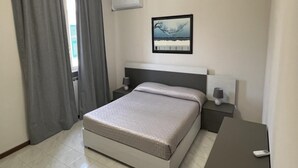 Una habitació d'hotel amb un llit, taules de nit, un aparell d'aire condicionat, un quadre a la paret i una finestra amb cortines.