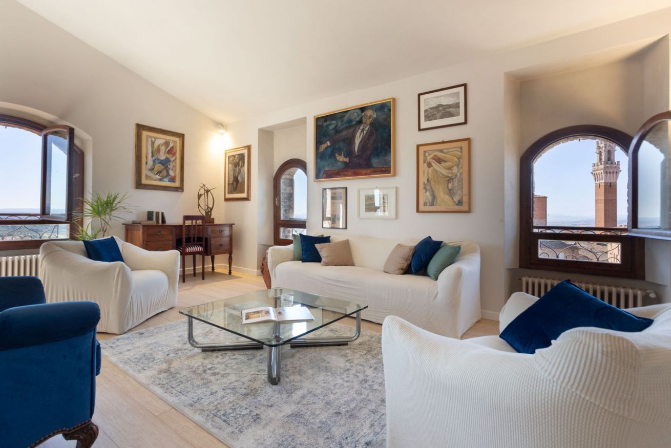 Un soggiorno spazioso con un divano bianco, poltrone blu e un tavolino in vetro. Alle pareti si notano opere d'arte incorniciate e una vista su una torre attraverso una finestra ad arco.