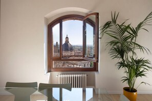 Une pièce dotée d’une grande fenêtre en arc de cercle donnant sur un paysage urbain, d’un radiateur en dessous, d’une table en verre avec des chaises et d’une plante en pot.