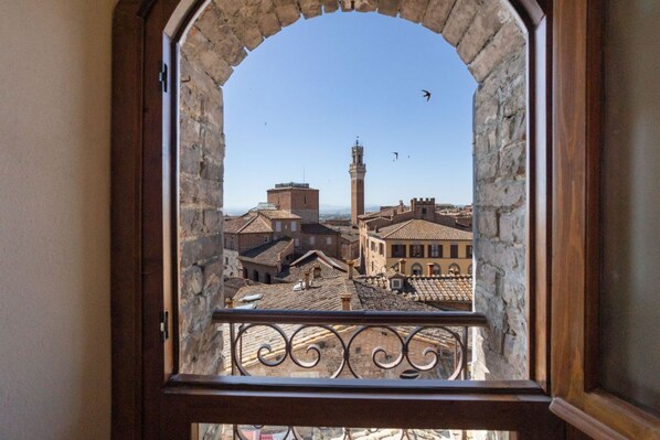 A view through a stone-framed window with a wrought iron balcony, overlooking a historic town with traditional buildings and a prominent tower.