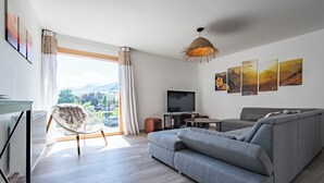 A modern living room with a grey sofa, a round coffee table, a TV, and a view of mountains through the window.