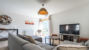 A modern living room with a grey sofa, a round coffee table, and a flat-screen TV. There is a wicker pendant light, a white radiator, and a view of the outdoors through a window.