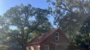 A small log cabin with a metal roof, surrounded by trees and a gravel driveway. - Fun Cabin in the Woods (Longdale)