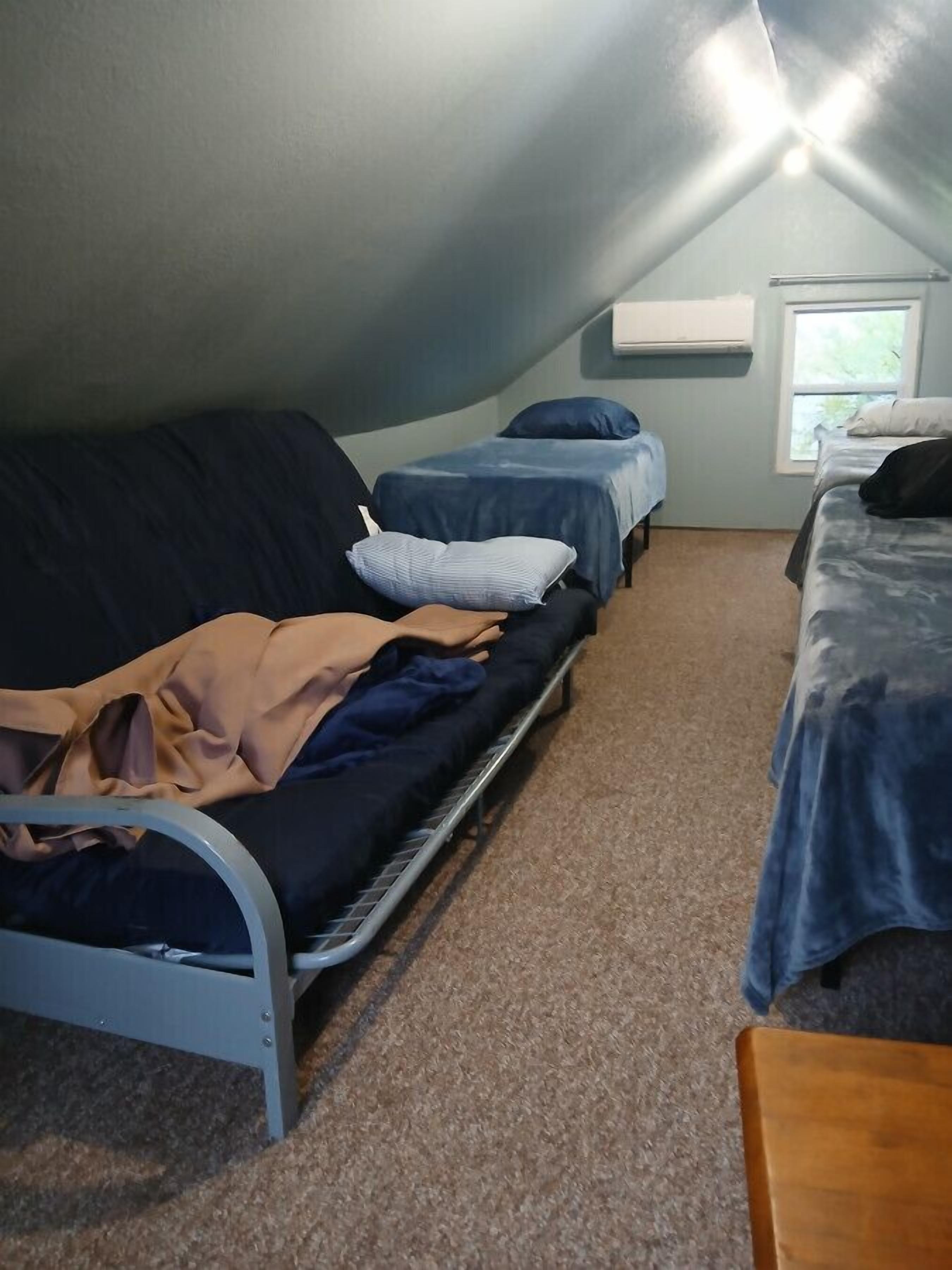 Ein kleiner, schwach beleuchteter Raum mit schrägem Dach, einem Bett, einer Couch und einem Fenster.