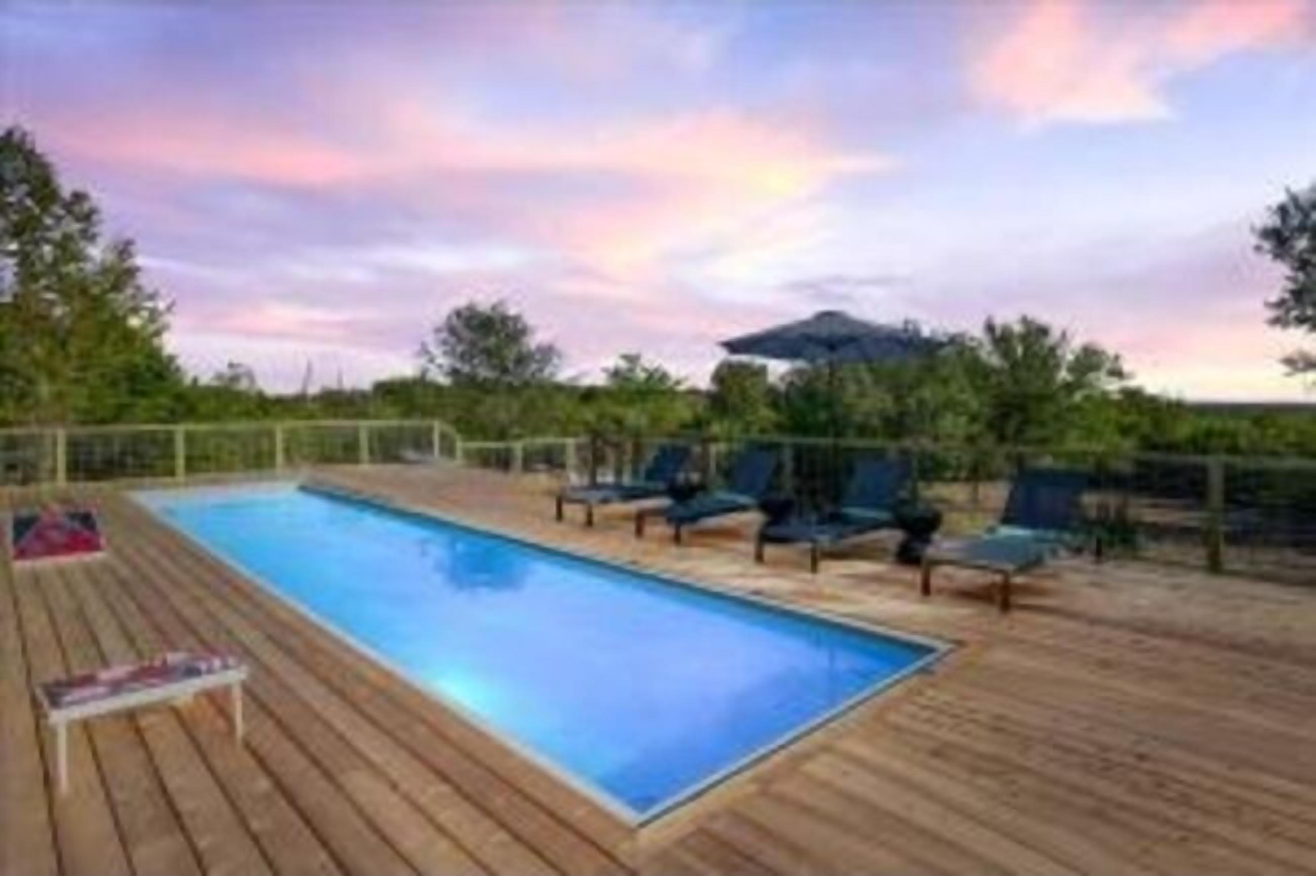 A rectangular swimming pool with a wooden deck, surrounded by lounge chairs and a covered area.