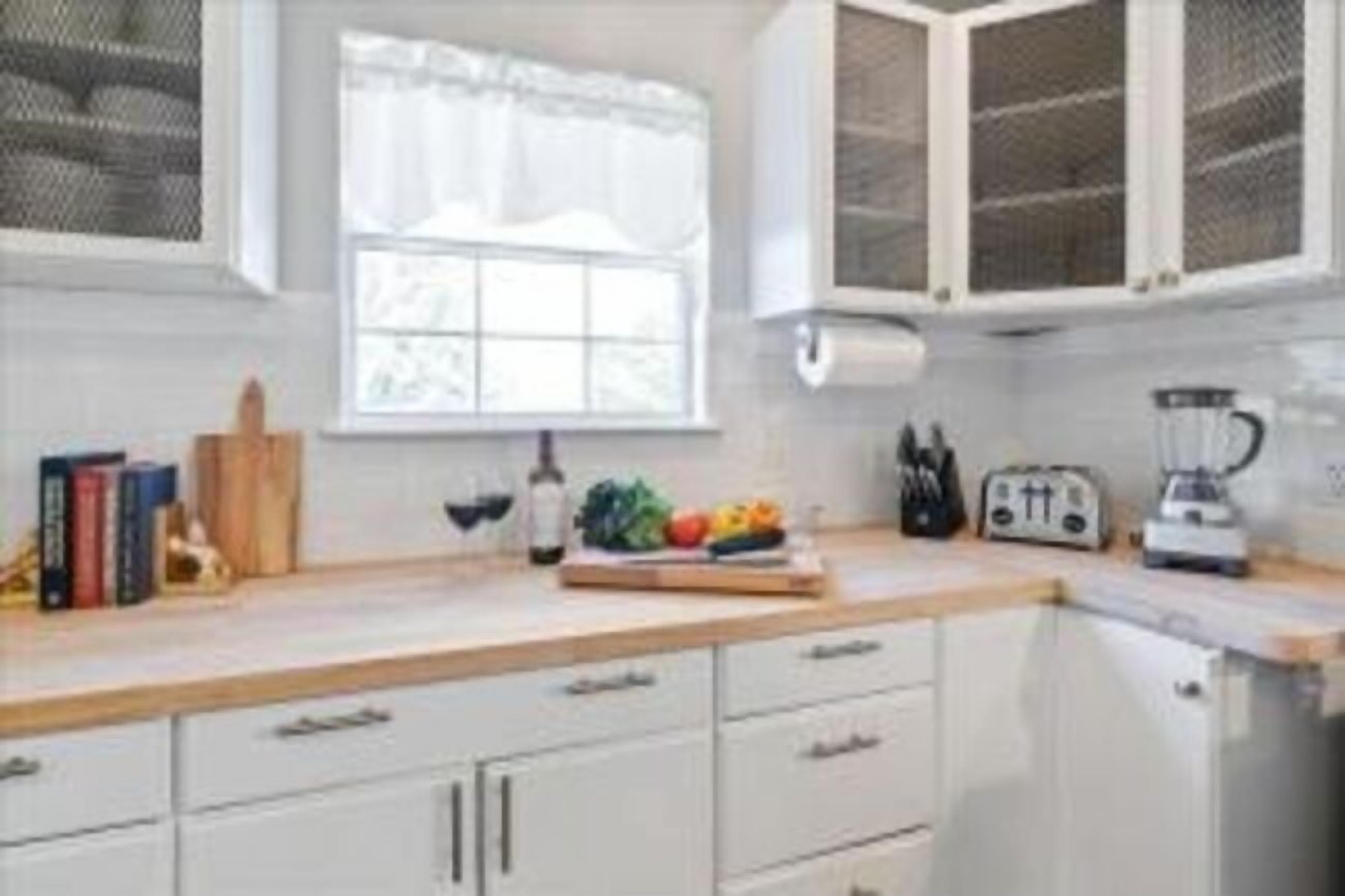 A kitchen with white cabinets, a window with white curtains, a fruit bowl, a toaster, and a blender.