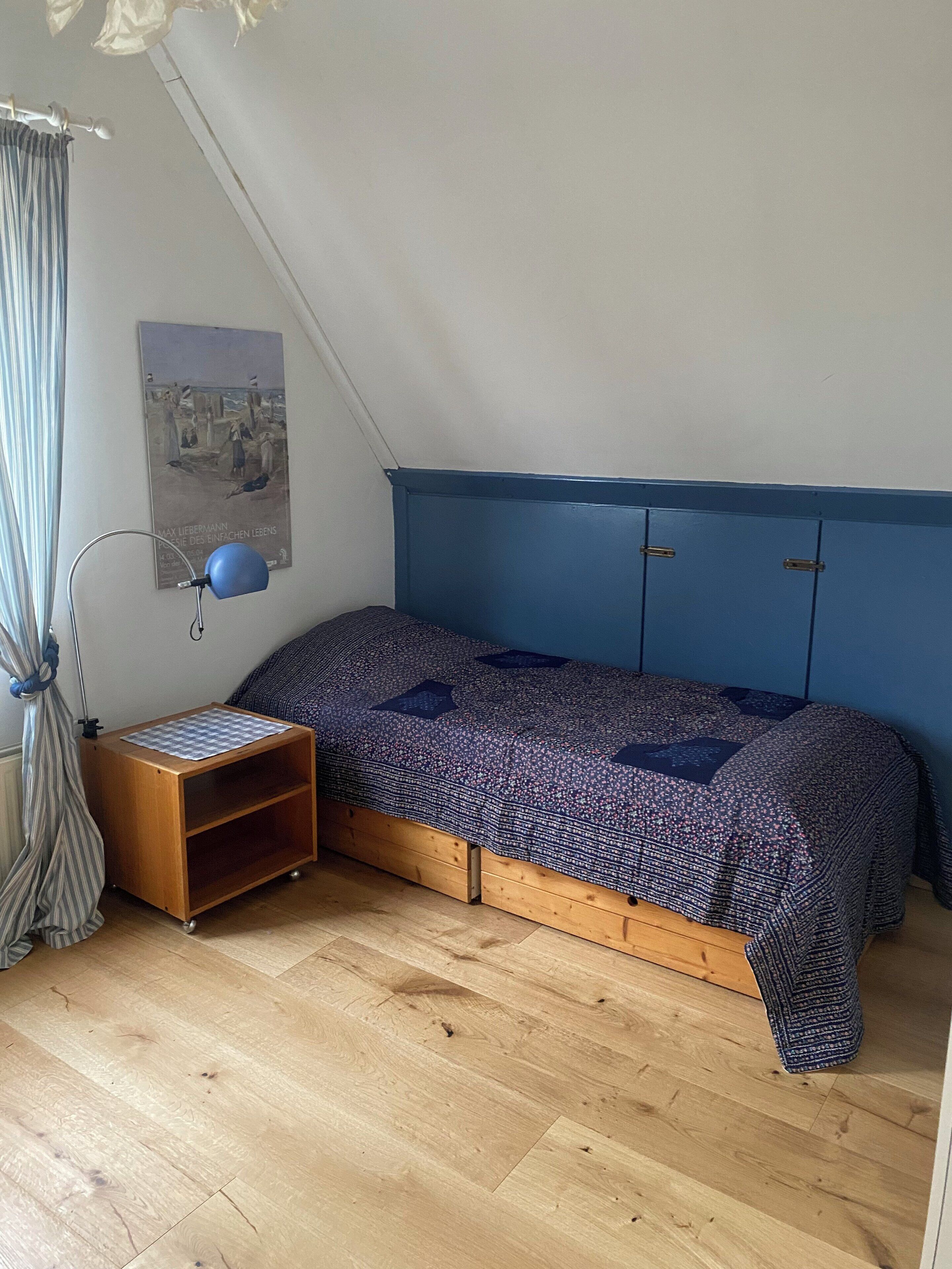 A bedroom with a sloped ceiling, a bed with a patterned bedspread, a small wooden nightstand, a blue lamp, and a poster on the wall.